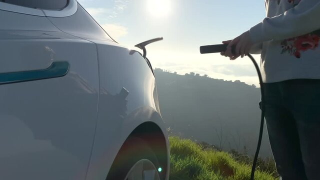 Slow motion of woman plugging in power cable to zero emission electric car at charging station with nature landscape. Cinematic 4K environmental and eco-friendly technologies, green, renewable concept