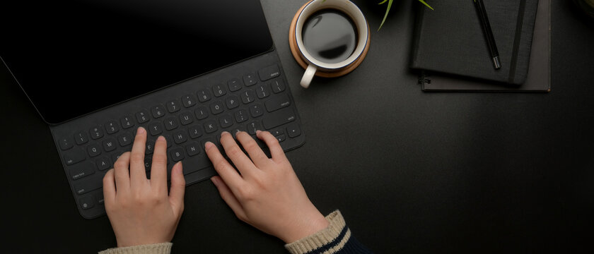 Hands typing on digital tablet keyboard on dark modern office desk with coffee cup and schedule books