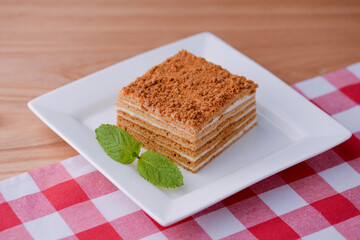 A slice of layered vanilla honey cake on a white plate with a mint leaf on wooden table with a plaid table cloth