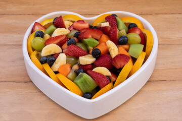 Fresh fruit salad in a white ceramic plate heart shaped. Kiwi, banana, strawberry, papaya, blueberry. Wooden background