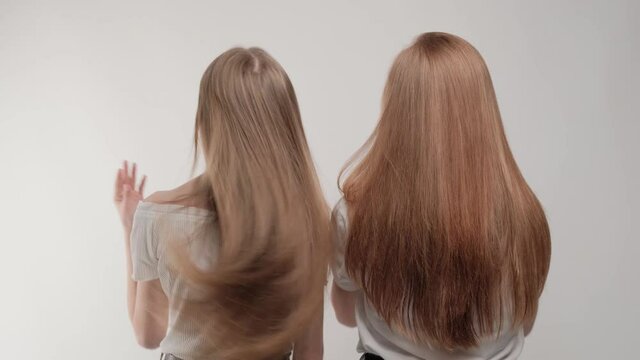 Blonde and red-haired girls stand with their backs and hair develop. Studio gray background.
