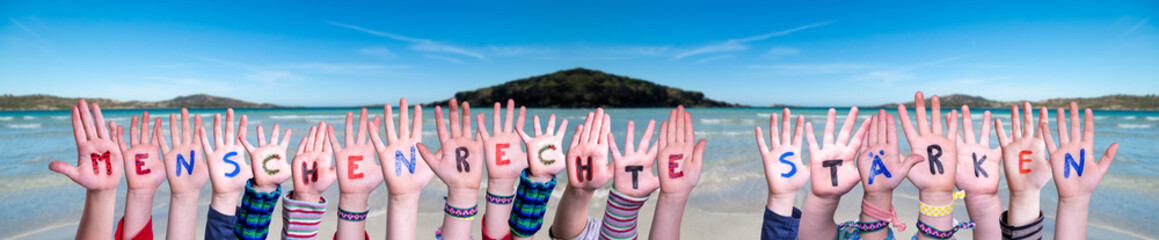 Children Hands Building Colorful German Word Menschenrechte Staerken Means Strengthen Human Rights. Ocean And Beach As Background