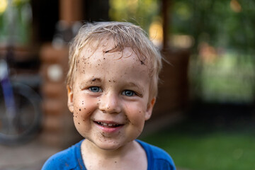Cute adorable blond caucasian little happy toddler boy portrait with messy mus spots on face after playing watering garden at yard or countyside farm. Childhood happiness summertime country concept