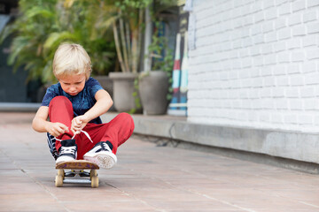 Little skateboarder sit on skateboard tying shoelaces before children training class in park. Active family lifestyle, outdoor activities on summer holidays in city. Kids sports, urban fashion