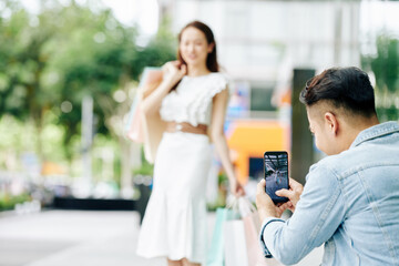Young man taking photo of girlfriend posing outdoors with shopping bags