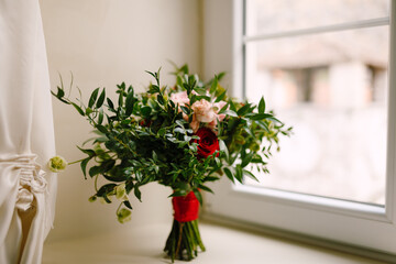 Fototapeta na wymiar bridal bouquet of red and pink roses, boxwood branches, not blooming buds of white flowers and red ribbons on the window