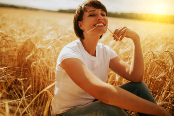 a smiling woman sitting in a wheat field under a sunset