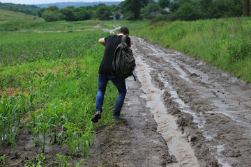 A young man with a backpack is trying to walk on a wet dirt road in heavy rain. Travel difficulties - obrazy, fototapety, plakaty