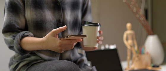 Female worker sitting on worktable and using smartphone while holding coffee in office room