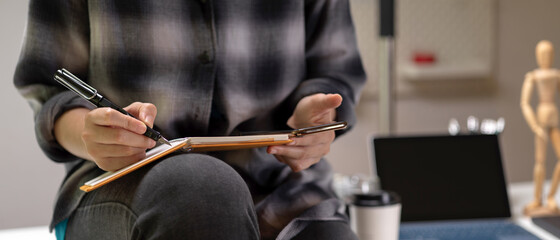 Female worker sitting on worktable and taking note on schedule book while looking on smartphone