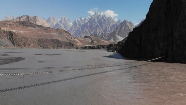 Forward-flying view of the Hussaini suspension bridge over the Hunza river with the Passu Cones in the background, snowy peaks and blue sky, Gilgit Baltistan region
