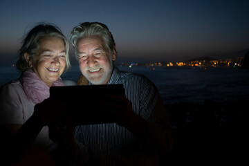 Cheerful senior couple using digit tablet sitting outdoor at night. Palm tree and night lights on background - concept of happy retired people tech and social