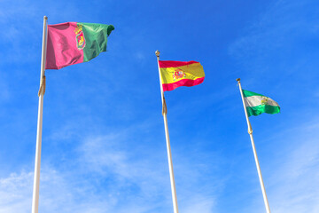 Flags of Andalusia, Granada and Spain, waving in a cloudy sky. Free sky background for copy space and sun shining.