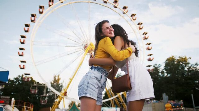 Two young women are smiling, hugging each other, talking and rejoicing their meeting while posing in park against a ferris wheel. Slow motion