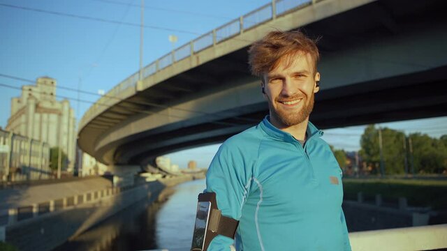 Handsome young athlete leaning on bridge railing while catching breath and recovering after jogging workout. Bearded man turning head to camera and smiling, headphones in his ears