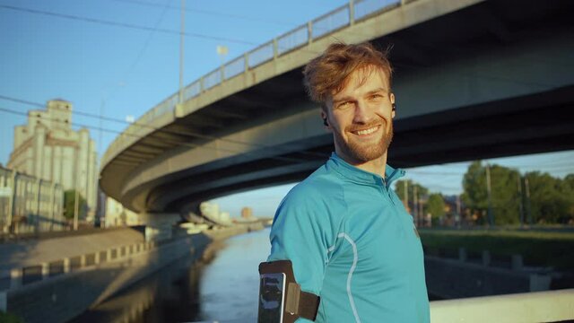 Handsome young athlete turning to camera and smiling happily while resting after jogging on bridge over river, headphones in his ears