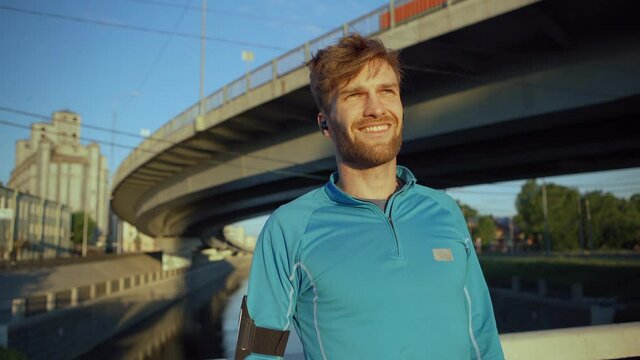 Handsome young athlete looking away dreamily, sighing and smiling while resting after jogging workout standing on bridge over river in evening light