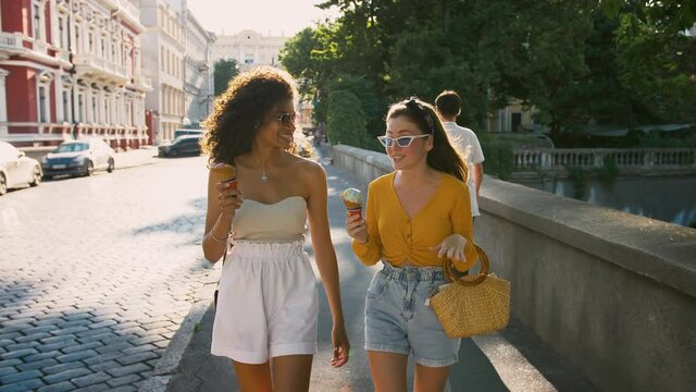 Two young females in casual outfit and sunglasses are eating ice-cream, talking and smiling while walking down the city street. Slow motion
