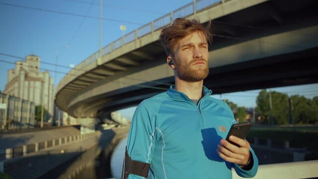 Handsome young man getting text message on cell phone while resting after jogging. Athlete reading sms and thinking standing on bridge over river