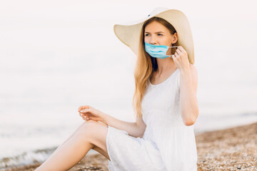 Fototapeta na wymiar attractive girl in a summer hat, with a medical protective mask on her face, poses on the beach, sitting on the sand by the sea. Summer vacation, coronavirus, quarantine