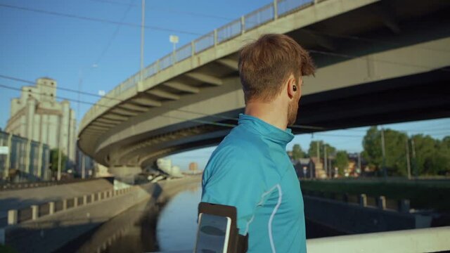 Waist up portrait of handsome male athlete turning his head and smiling happily at camera while standing on bridge over river after jogging