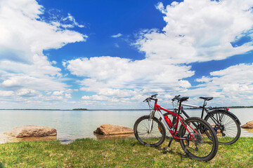 Two bikes are on the picturesque background  with blue sky, white clouds, water and green grass.