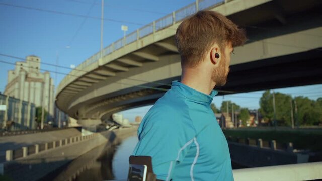 Waist up portrait of handsome male athlete looking around, checking time on smartwatch and smiling while standing on bridge over river after jogging