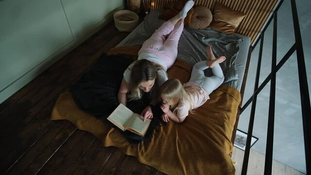 Adult mother with girl in pink white pajama laying on bed reading book upview