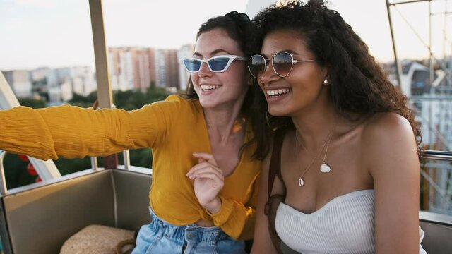 Young girls in sunglasses and casual outfits are smiling, talking and looking surprised while sitting in a ferris wheel booth. Close-up, slow motion