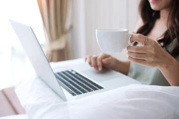 casual woman holding coffee cup while using computer laptop on pillow in bedroom. Young adult female Working from home or online shopping in morning. Freelancer, lifestyle and technology concepts