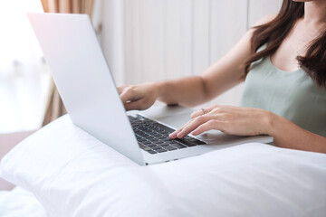Happy casual woman sitting and using computer laptop on white pillow in bedroom. Young adult female Working from home or online shopping in morning. Freelancer, lifestyle and technology concepts