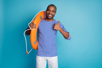 Portrait of positive afro american guy hold circle lifeguard buoy approve life sand safety quality show thumb up sign wear sailor striped shirt white shorts isolated blue color background - Powered by Adobe