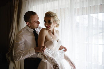 Gorgeous, blonde bride in a white luxurious dress getting ready for the wedding. Morning preparations. A woman puts on a dress. The groom helps the bride.