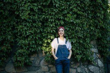 Slim sexy brunette girl wearing woman's denim overall over green tree wall texture in urban area