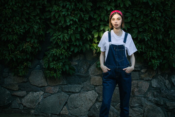 Slim sexy brunette girl wearing woman's denim overall over green tree wall texture in urban area