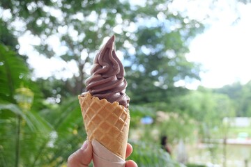 In selective focus a female hand holding a cone of chocolate ice cream with blurred green bokeh and nature background in a garden area
