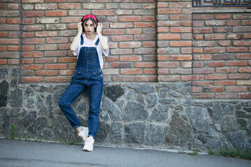 Slim sexy brunette girl wearing woman's denim overall over green tree wall texture in urban area