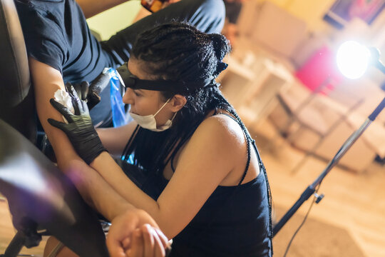 Tattoo Artist On Action, Doing A Tattoo On Customer's Arm, Wide Angle Shot, Artist Is Recognizable, Wearing A Face Mask