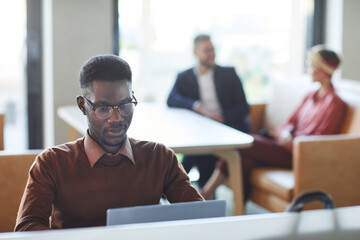 Portrait of young African-American man dressed in business casual using laptop in office or coworking space with people in background, copy space