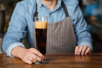 Dark ale with foam. Barman holds high glass of beer on wooden bar counter