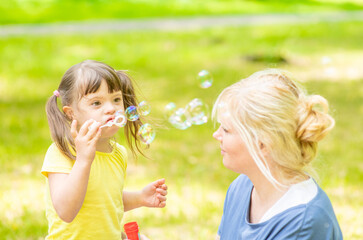 Happy little girl with syndrome down blows bubbles in a summer park with her mother