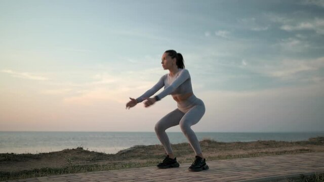 Young sexy caucasian woman making squat jumps by the sea. Outdoor workout on sunset. Athlete female exercising. Long shot