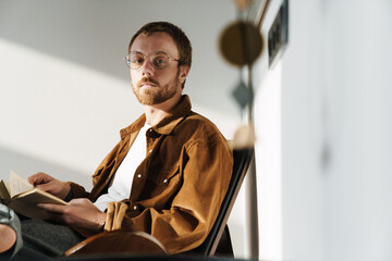 Photo of thinking young man reading book while sitting in armchair