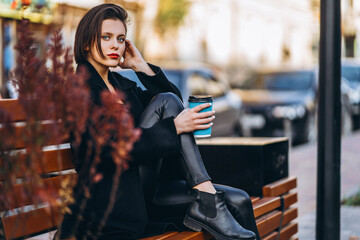 Fototapeta premium Young woman with short haircut, dressed in black, sits on a bench in the city. A woman holds a cup of coffee and looks into the camera