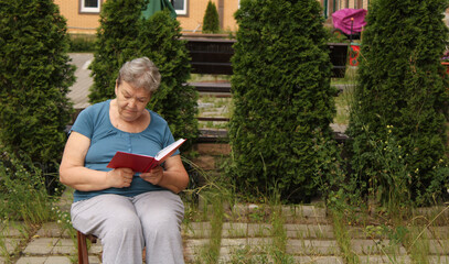 An elderly woman is sitting in the courtyard on a summer day and reading a book. The concept of a happy old age and International Day of Older Persons and Grandparents' Day