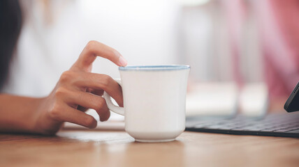 Closeup image of woman’s hand holding white mug while sitting at office desk with tablet computer.