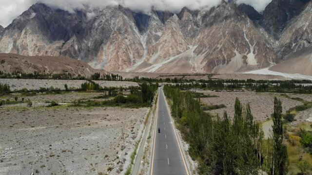 Epic aerial view of a man riding his motorbike toward the Passu Cones, on the KKH Karakorum Highway, crossing a green valley, peaks and mountains during a foggy day, Pakistan