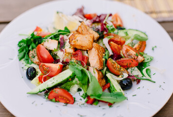 White plate with fresh vegetable salad with slices of grilled salmon on a wooden table on the terrace.