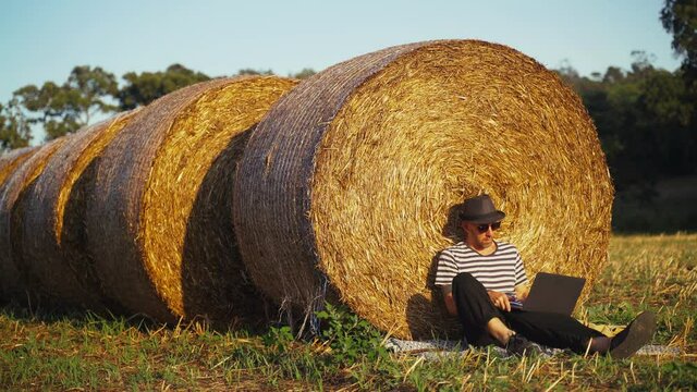 A young man works with laptop in the field. A man in a stripe tshirt and a black hat sitting leaning on a haystack and typing on a laptop keyboard. Vacation in the village. Sunny summer day. Holidays