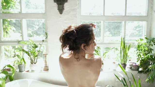 Sexy woman posing near the window and bathtub with naked back. Looking at the camera. Slowmotion.